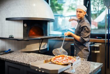 Chef puts freshly baked hot pizza from a spatula in a box. Catering kitchen work.