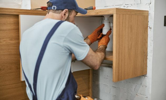 Male plumber in cap repairing pipe in kitchen