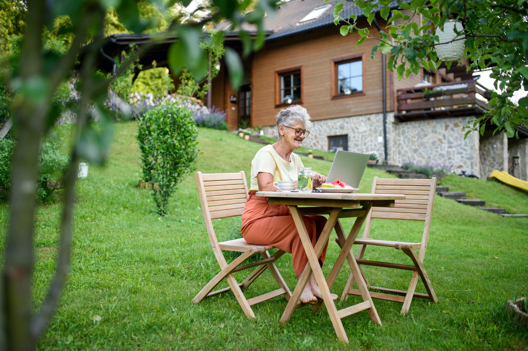 Happy senior woman with laptop working outdoors in garden, home office concept