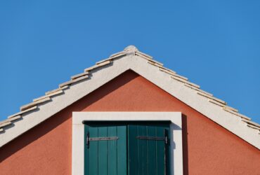 The house on the blue sky background. Roof and window with shutters.