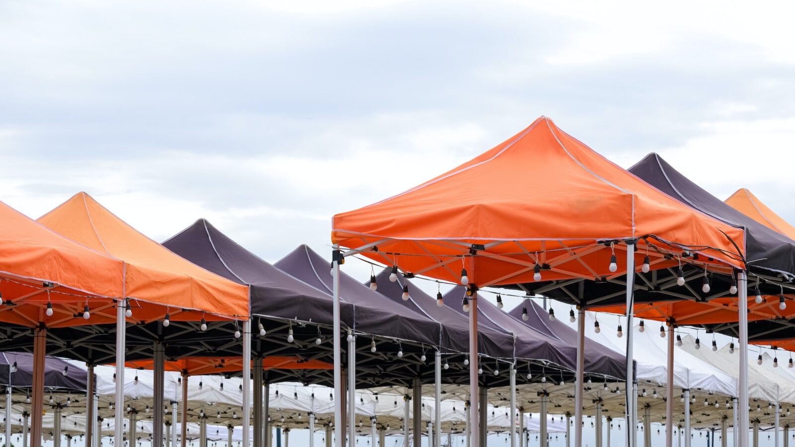 Many foldable canopy tents with light bulbs decoration for outdoors event against cloudy sky