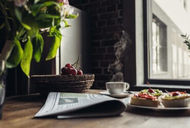 Still life of newspaper, breakfast with cakes and hot coffee on kitchen table in front of window