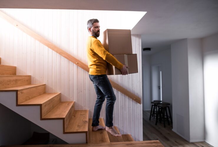 Mature man walking down stairs in house, holding moving boxes.