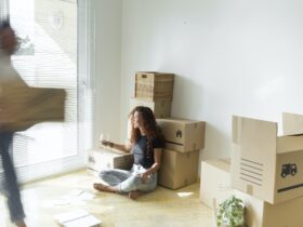 Young woman sitting in new home with glass of red wine while friend is moving fast with cardboard bo