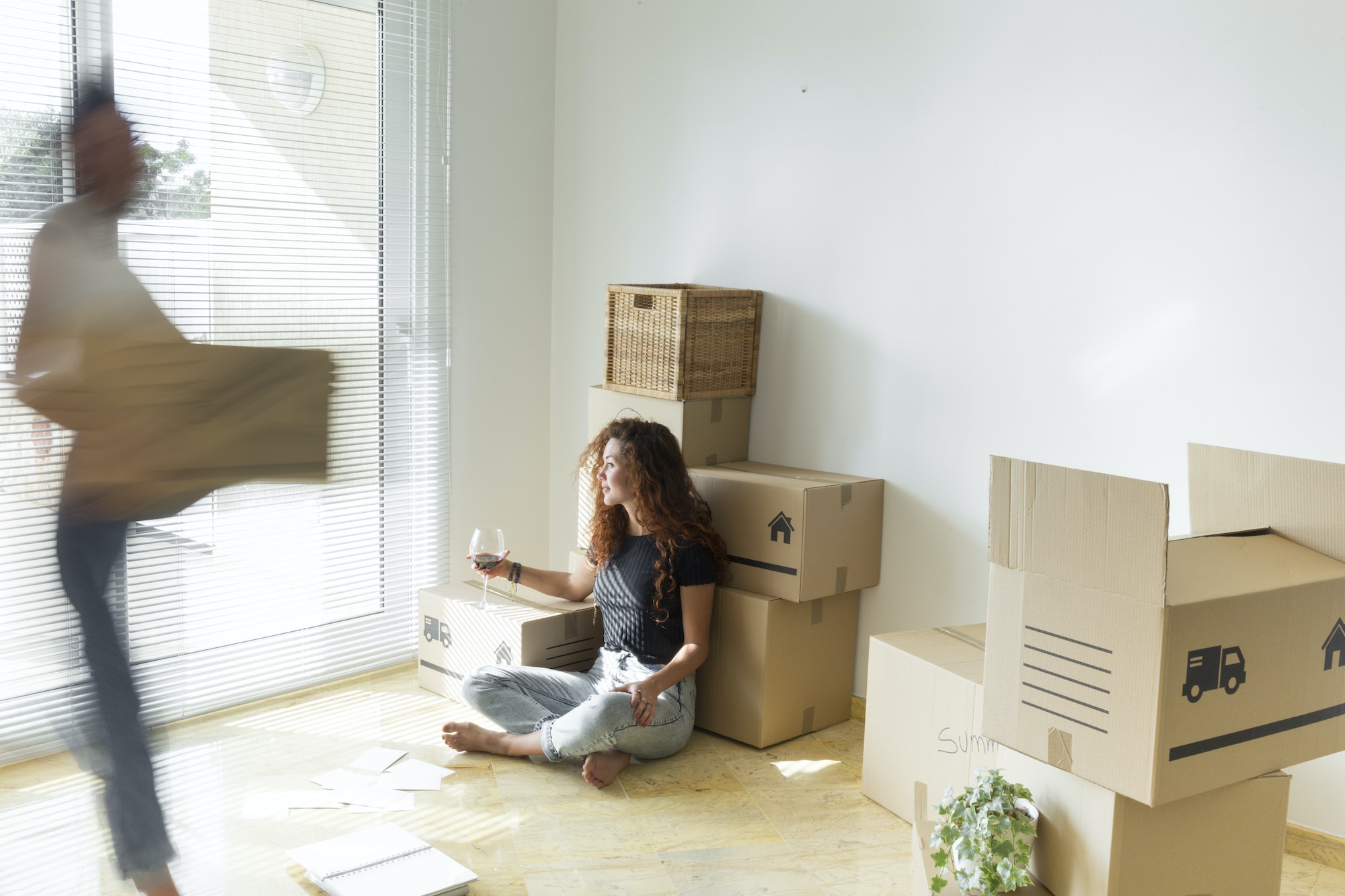 Young woman sitting in new home with glass of red wine while friend is moving fast with cardboard bo