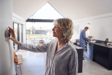 Mature Woman Adjusting Wall Mounted Digital Central Heating Thermostat Control At Home