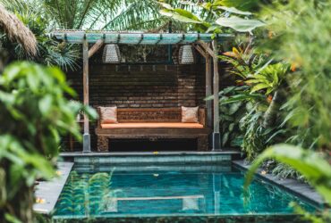 Small private swimming pool in Bali house. Green tropical plants around, wooden sofa.