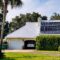 Home with beach vibes has solar panels on rooftop surrounded by tropical palm trees.
