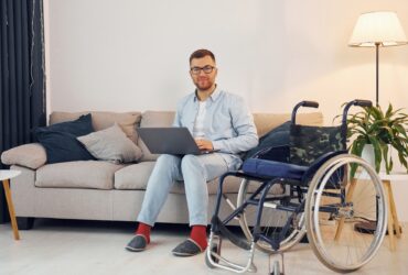With laptop on the legs. Disabled man in wheelchair is at home
