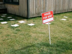 A red 'House for Rent' sign stands on a grassy lawn beside a wooden house exterior.