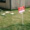 A red 'House for Rent' sign stands on a grassy lawn beside a wooden house exterior.