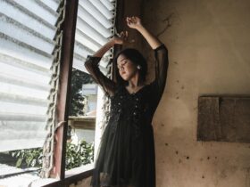 A serene portrait of a woman in a black dress with arms raised, standing by a window.