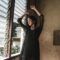 A serene portrait of a woman in a black dress with arms raised, standing by a window.