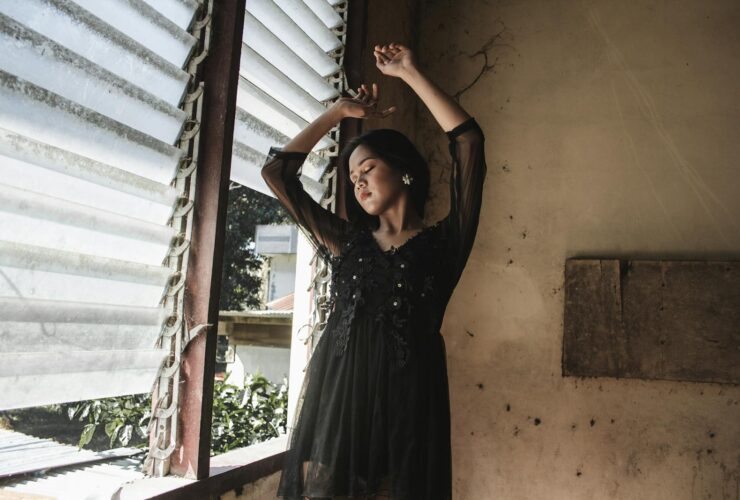 A serene portrait of a woman in a black dress with arms raised, standing by a window.
