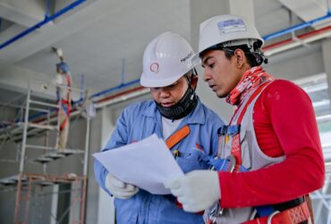 Two engineers in safety helmets reviewing construction plans at a worksite.