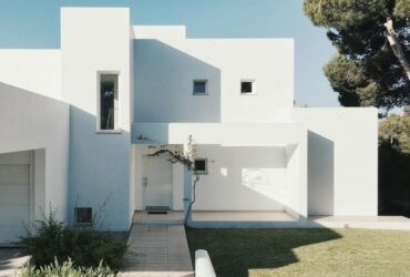 A minimalist modern white house exterior with clean lines and geometric shapes, set against a clear blue sky.