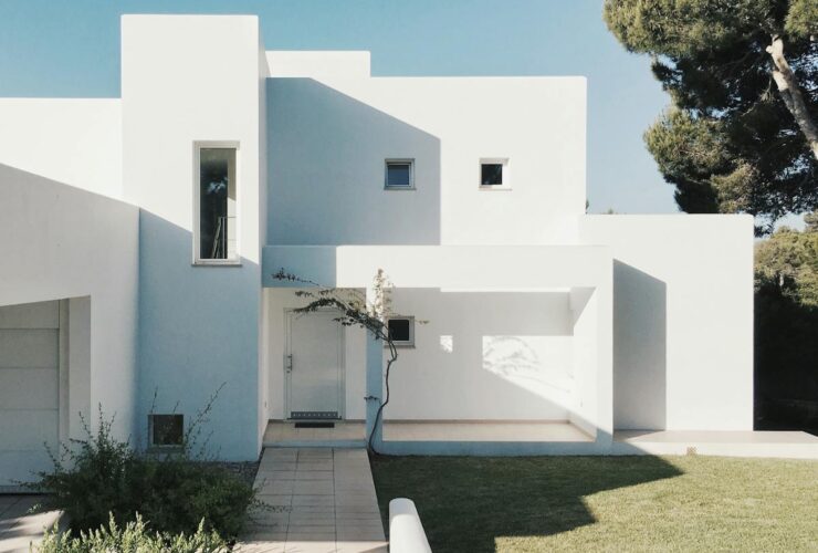 A minimalist modern white house exterior with clean lines and geometric shapes, set against a clear blue sky.