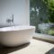 Sleek white bathtub by large window in sunlit minimalist bathroom setting.