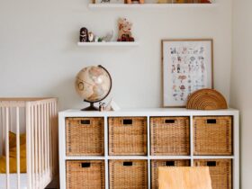 Comfortable bed with toys globe and paintings placed on wooden cabinet with wicker boxes and soft white rug with toy house on floor