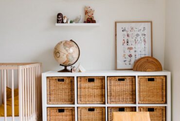 Comfortable bed with toys globe and paintings placed on wooden cabinet with wicker boxes and soft white rug with toy house on floor