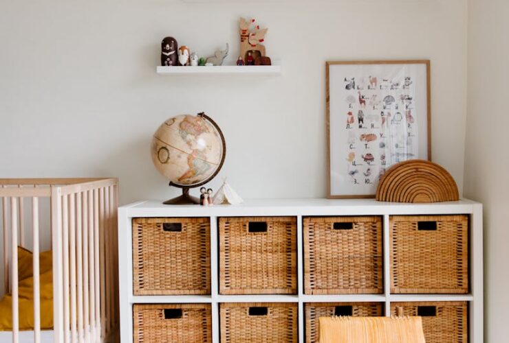 Comfortable bed with toys globe and paintings placed on wooden cabinet with wicker boxes and soft white rug with toy house on floor