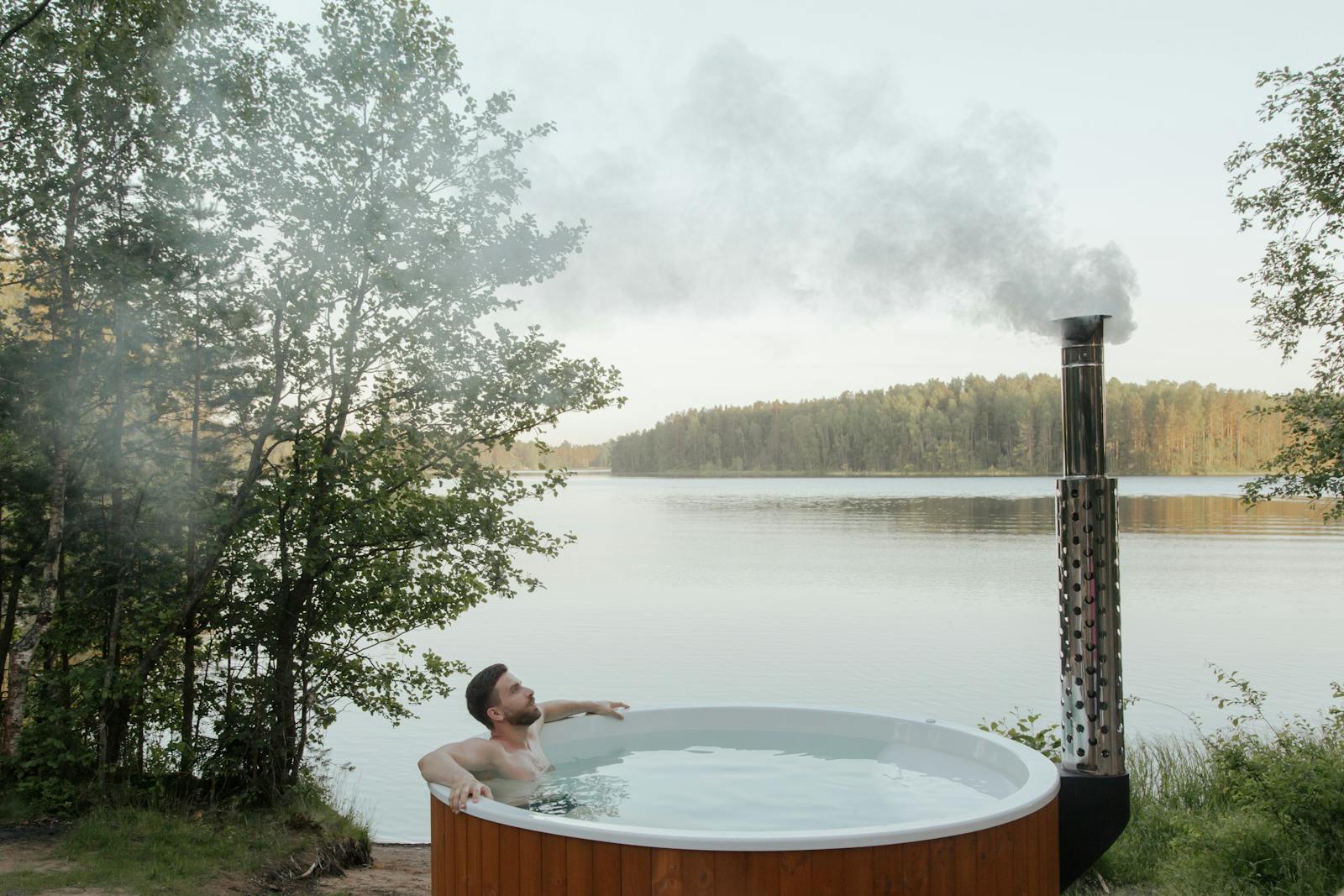 Man enjoying a serene soak in an outdoor hot tub by a peaceful lake surrounded by nature.