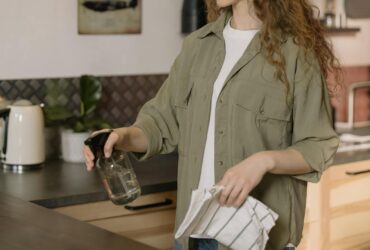 A woman with curly hair cleans a kitchen counter using a spray bottle and cloth.