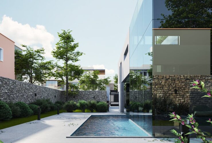 Elegant modern house with glass exterior and pool surrounded by lush greenery, under a clear sky.