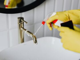 From above anonymous housekeeper in yellow rubber gloves spraying cleaner to chrome water faucet in bathroom
