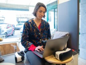 A young woman wearing a printed shirt works remotely on a laptop beside a VR headset in a cozy space.