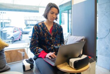A young woman wearing a printed shirt works remotely on a laptop beside a VR headset in a cozy space.