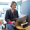 A young woman wearing a printed shirt works remotely on a laptop beside a VR headset in a cozy space.