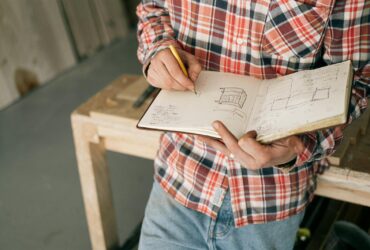From above of crop anonymous slim male carpenter in plaid shirt and blue jeans doing quick sketches of future furniture in notepad while leaning on workbench in workshop