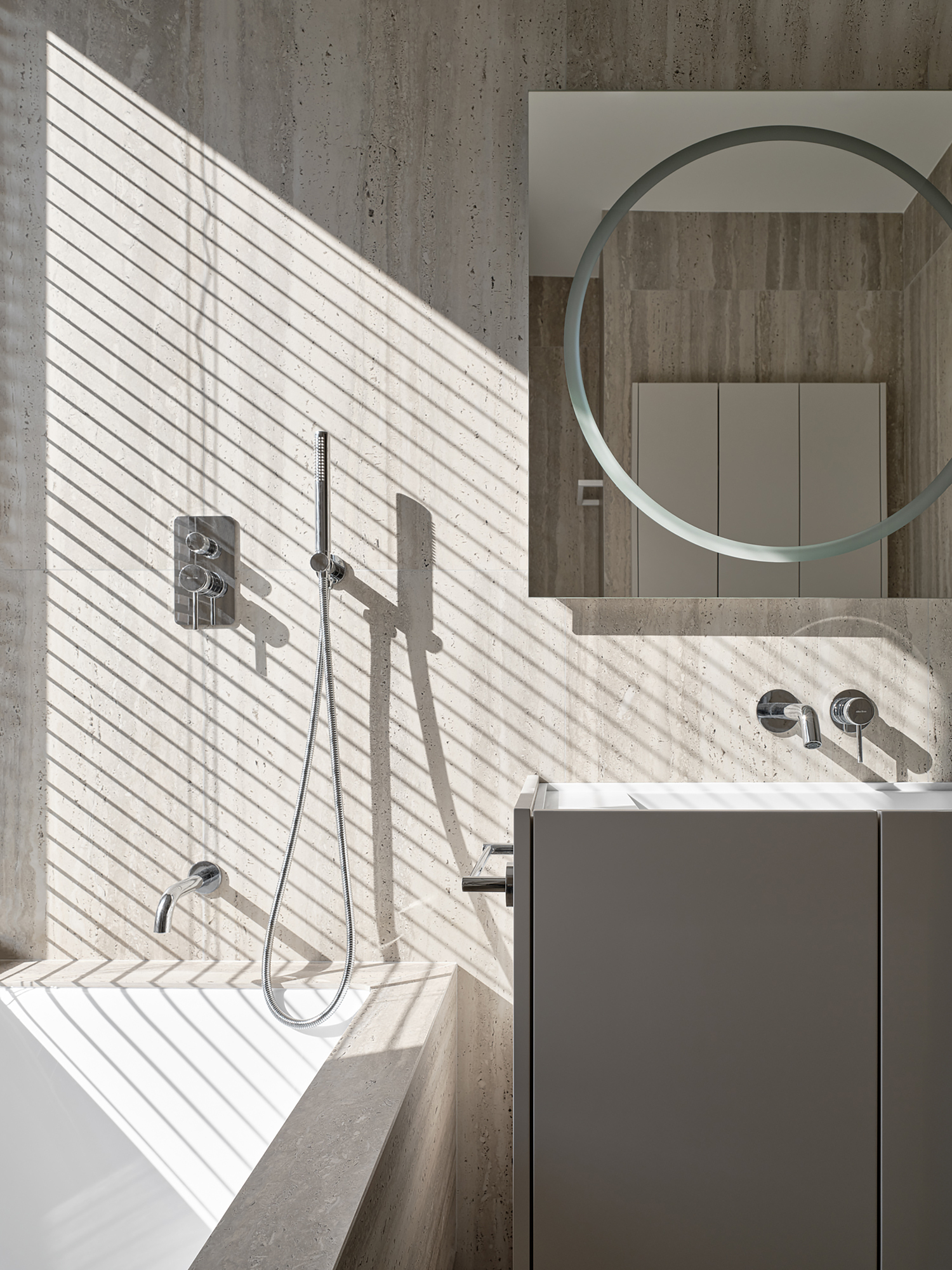 Master bathroom with travertine tiles, bathtub, round mirror, and bamboo blinds at window